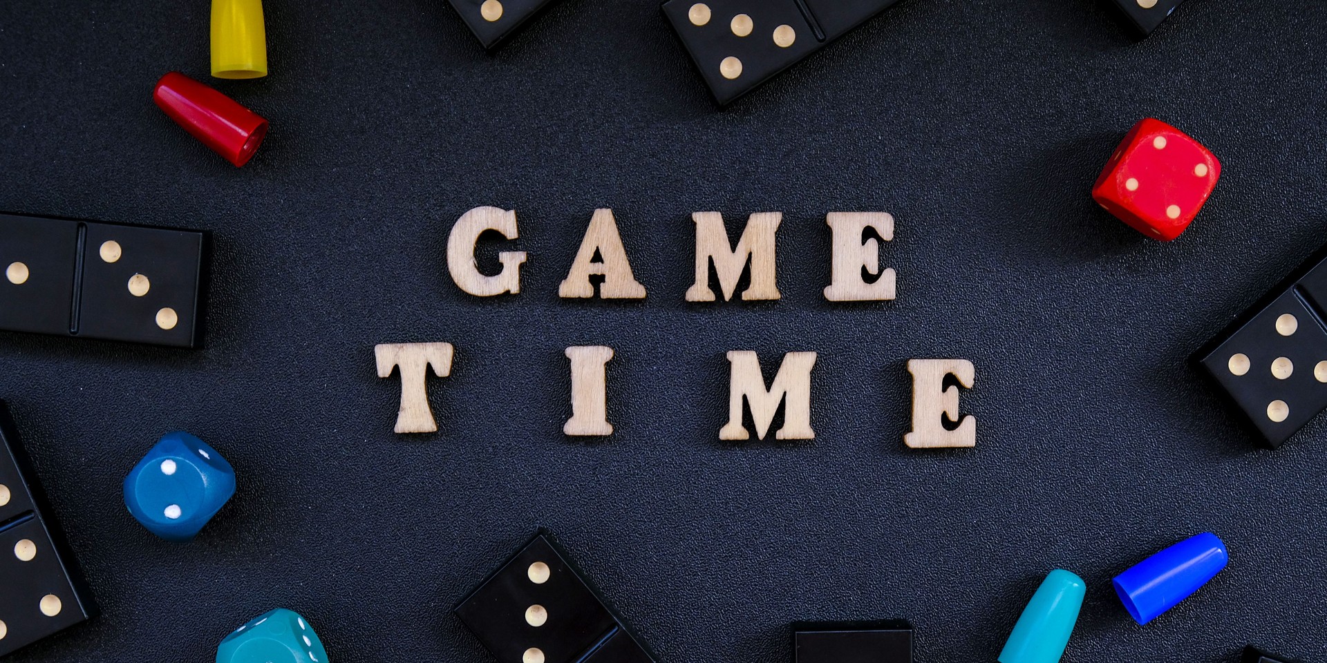 Text GAME TIME spelled out in wooden letter. Surrounded by dice, dominoes other game pieces on black background. Table games. Stay home activity